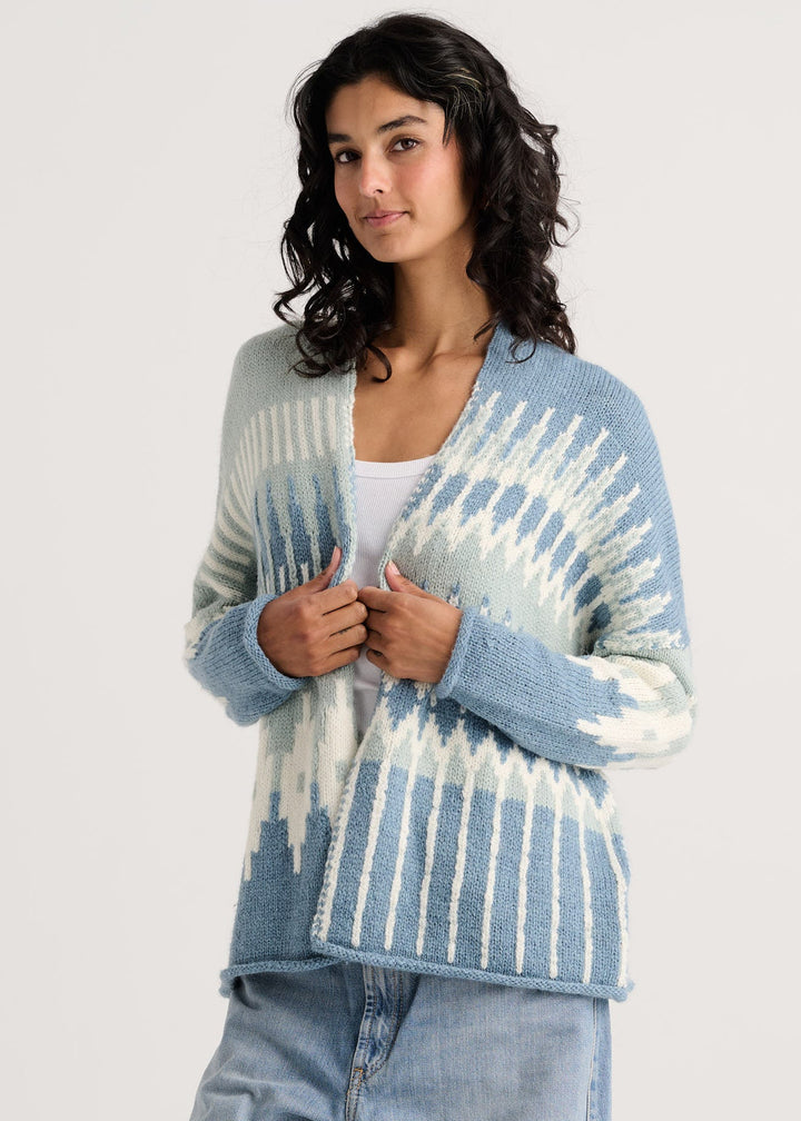 A woman wearing a light blue and white patterned knit cardigan over a white top and light blue jeans, photographed against a plain light gray studio background.