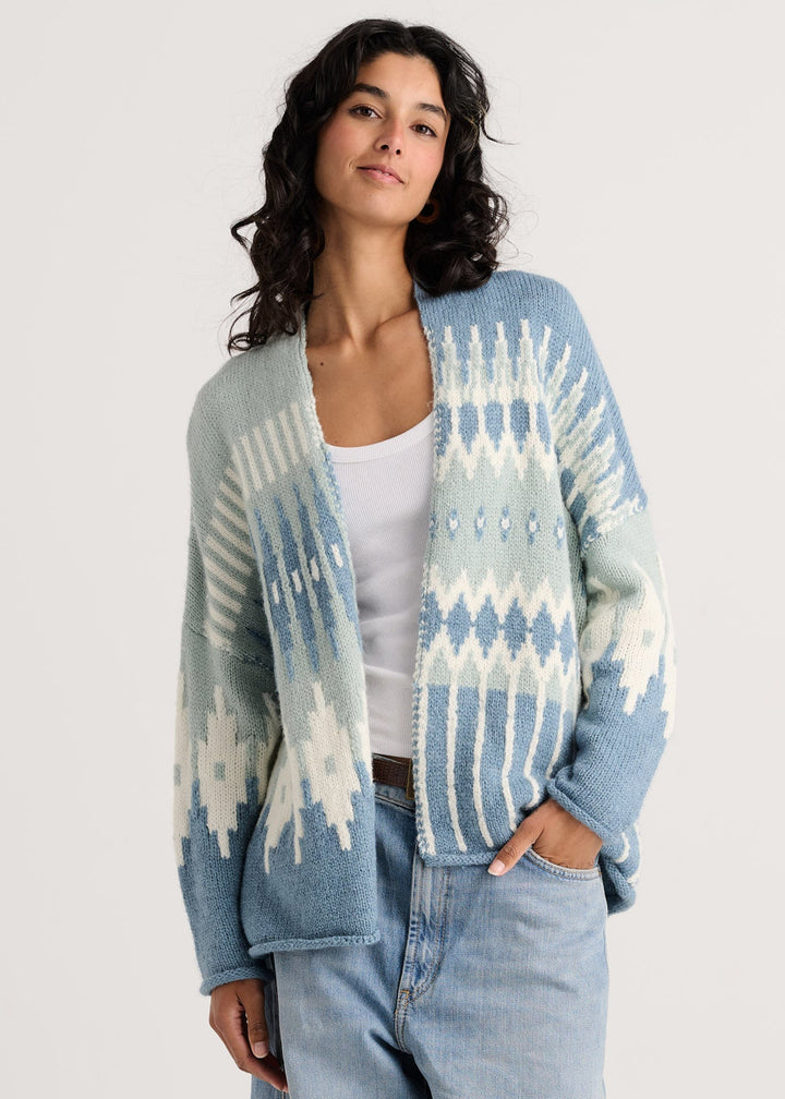 A woman wearing a light blue and white patterned knit cardigan over a white tank top and light-wash jeans, photographed against a plain light gray studio background.