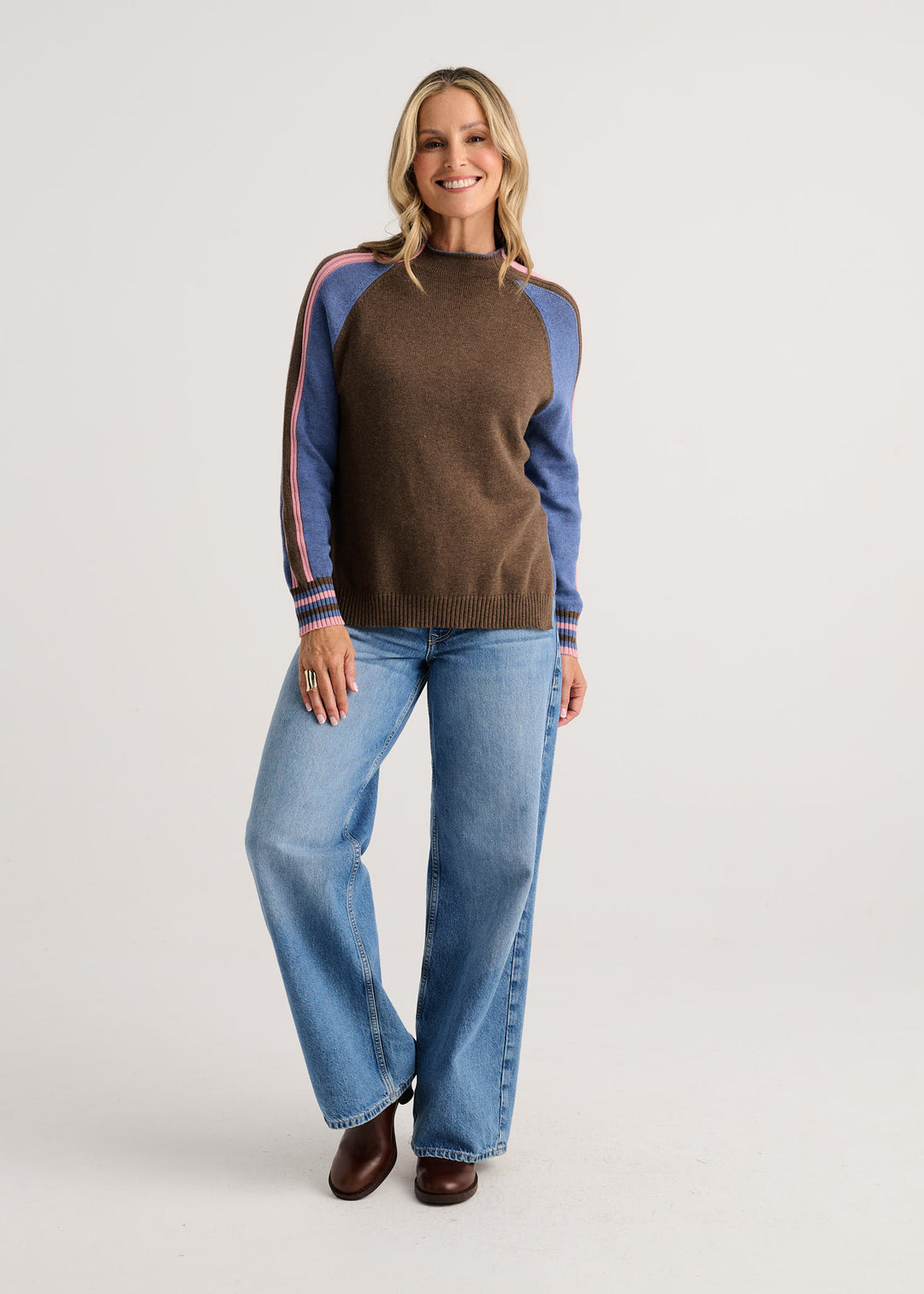 A woman modeling a brown knit sweater with blue raglan sleeves and striped cuffs, paired with blue jeans and brown boots, against a light gray studio background.