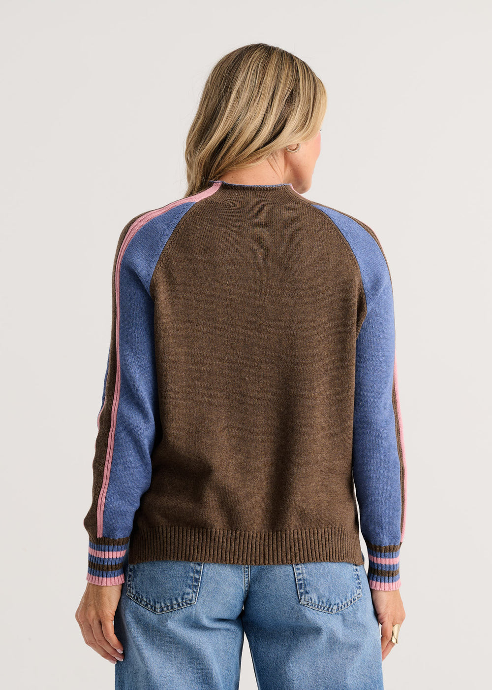 A woman viewed from the back wearing a brown knit sweater with blue raglan sleeves and pink stripe accents, paired with blue jeans, against a light gray studio background.