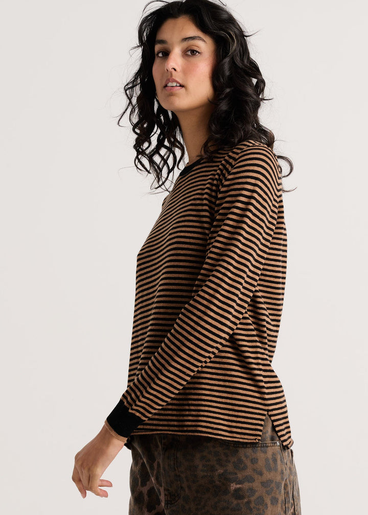 A woman with dark curly hair is shown in profile wearing a brown and black horizontally striped long-sleeve top and leopard-print pants against a plain light gray studio background.