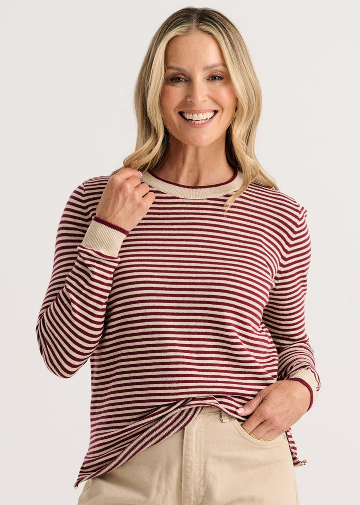 A smiling blonde woman wearing a long-sleeve knit top with horizontal burgundy and beige stripes, paired with light beige pants, against a plain light gray studio background.