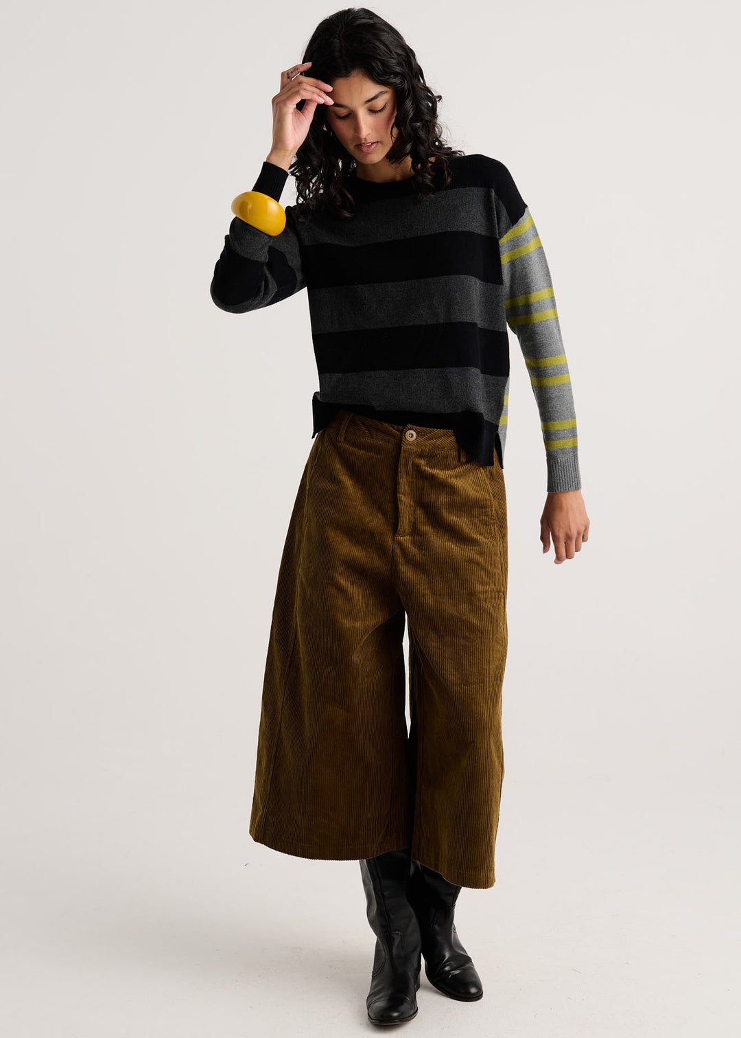 A model wearing a black and gray striped knit sweater with yellow and green striped sleeves, paired with brown wide-leg corduroy pants and black boots, against a light gray studio background.