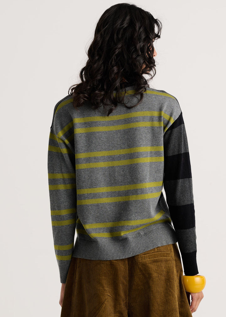A woman viewed from the back wearing a gray knit sweater with horizontal yellow stripes and black sleeve panels, paired with brown corduroy pants against a plain light background.