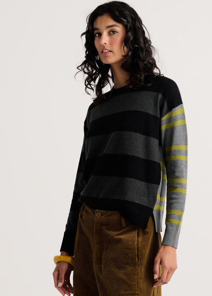A woman wearing a black and gray striped knit sweater with yellow-green striped sleeves, paired with brown corduroy pants, against a plain light gray studio background.