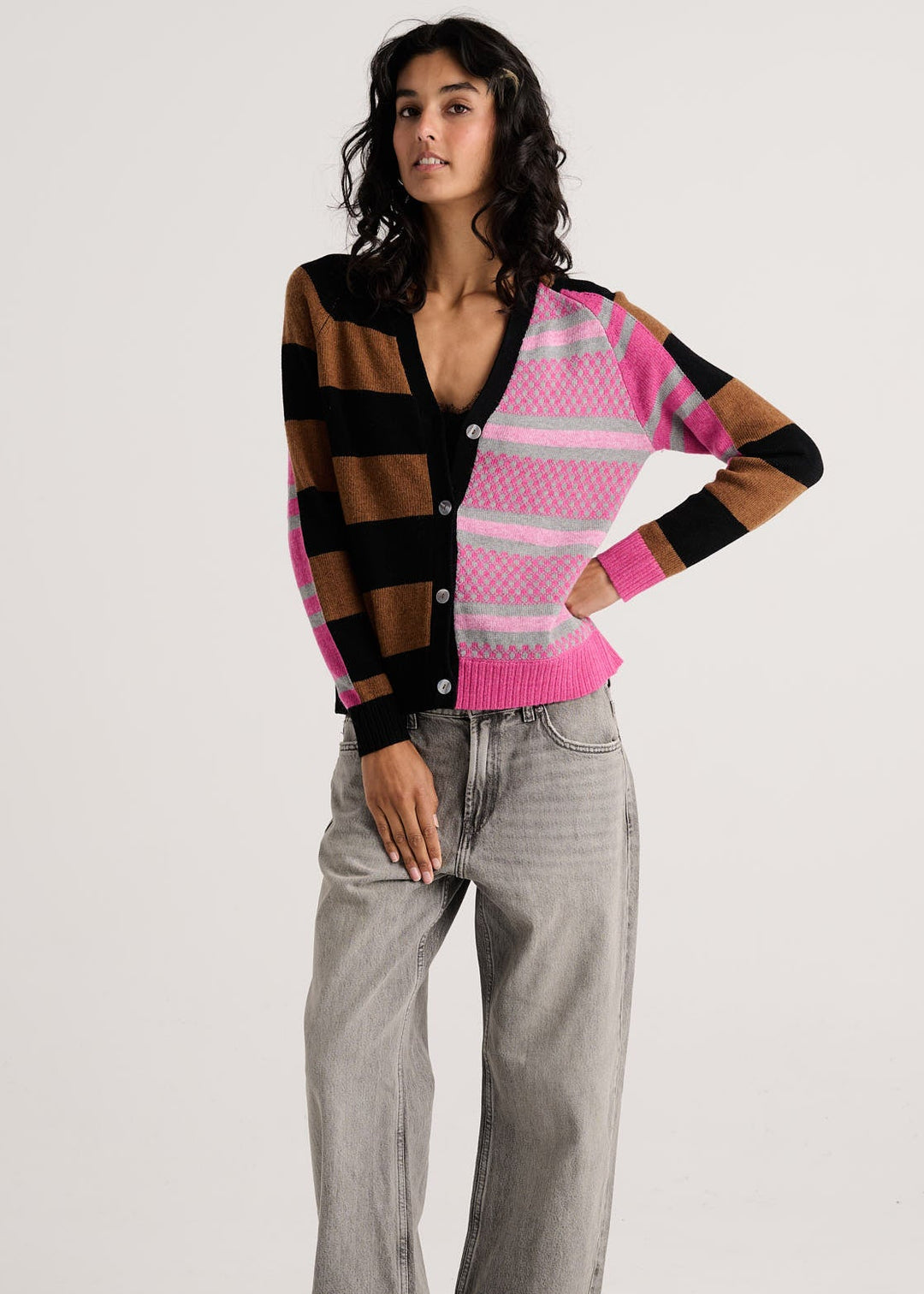 A model stands against a light gray studio background wearing a colorful knit cardigan with brown, black, pink, and white stripes, paired with light gray wide-leg jeans and brown shoes.