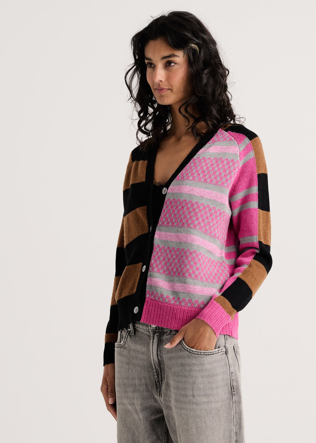 A woman wearing a colorful striped cardigan with pink, black, and brown blocks, paired with gray jeans, photographed against a plain light gray background.