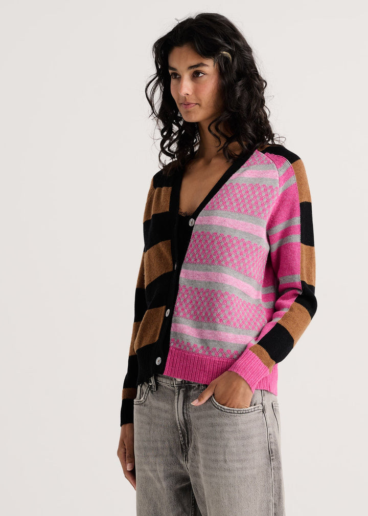 A woman wearing a colorful striped cardigan with pink, black, and brown blocks, paired with gray jeans, photographed against a plain light gray background.
