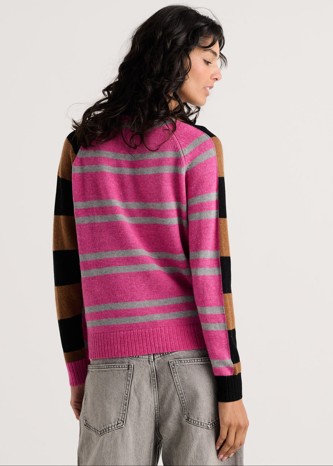 A woman shown from the back wearing a pink knit sweater with horizontal gray stripes and brown and black striped sleeves, paired with light gray jeans, against a plain light gray studio background.