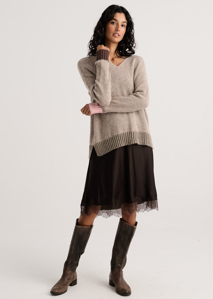 A model stands against a light gray studio background wearing a beige knit sweater, a dark brown pleated skirt with lace trim, and brown knee-high boots.