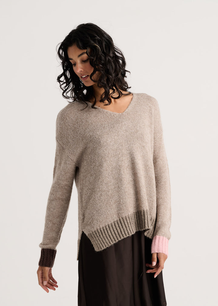 A woman wearing a light taupe knit sweater with contrasting dark brown ribbed cuffs and hem, layered over a dark skirt. The photo is taken against a clean, off-white studio background.