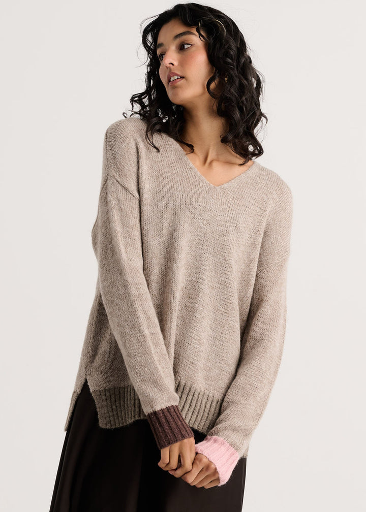 A woman wearing a light taupe-beige knit sweater with a V-neck and ribbed cuffs, paired with a dark skirt, photographed against a plain light gray studio background.