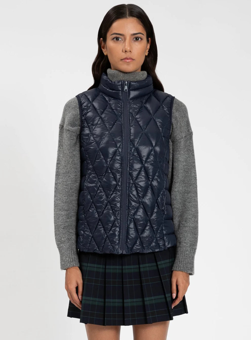 Woman wearing a navy quilted vest over a gray sweater with a plaid skirt on a white background