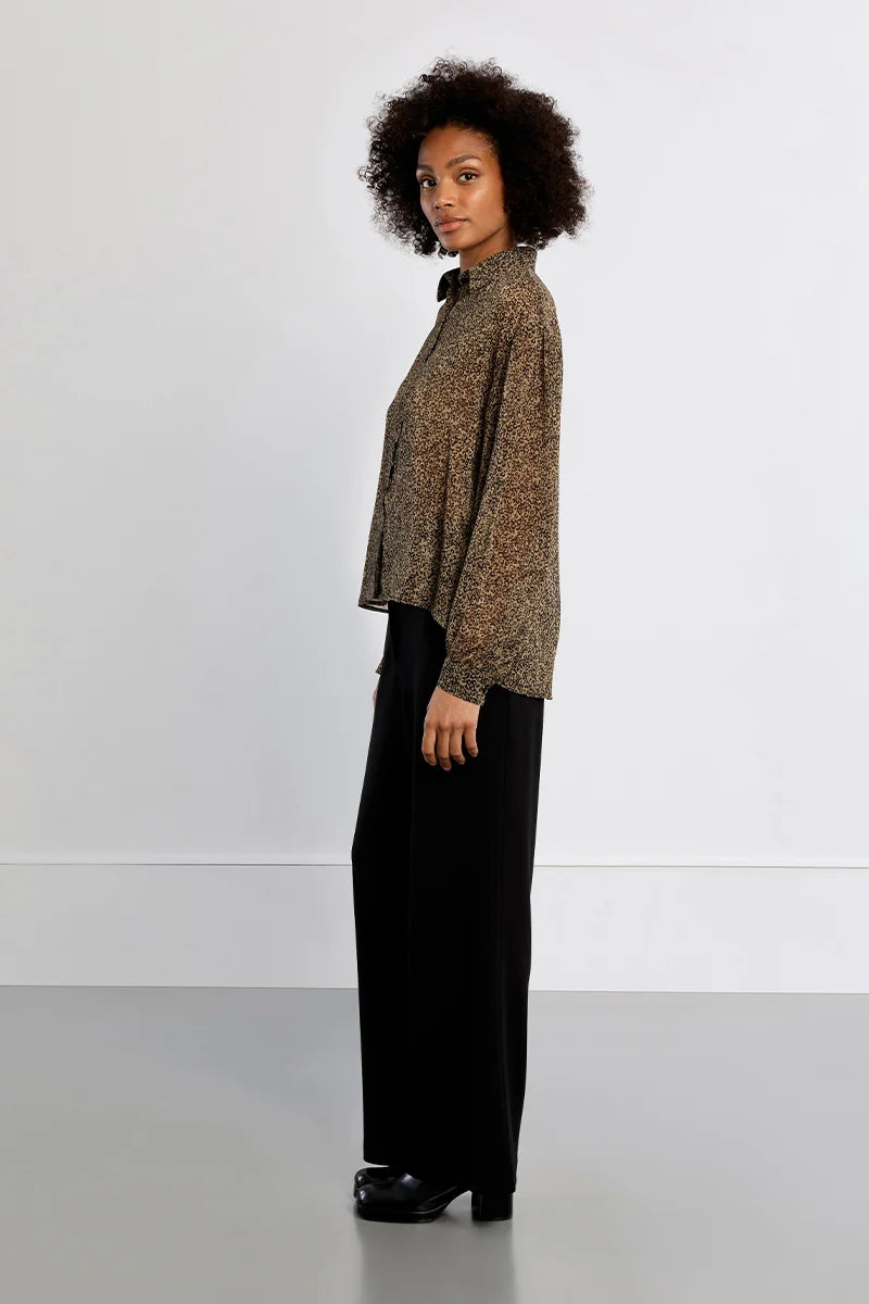 A woman with curly dark hair stands in profile wearing a loose, patterned brown and beige top with black wide-leg trousers and black shoes, photographed against a plain light gray studio background.