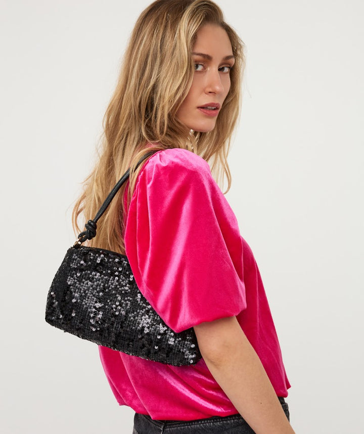 A woman wearing a hot pink velvet top and black jeans, carrying a black sequined shoulder bag with a black strap against a plain light gray background.