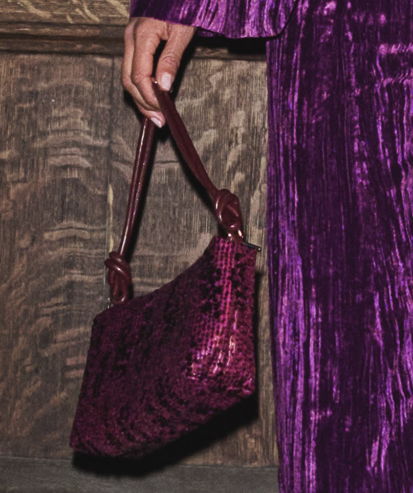 A close-up of a person holding a small structured handbag with a dark burgundy handle and a textured, sequined or crushed-velvet body in a deep plum-purple color. The person is wearing a matching purple velvet garment, standing against a rustic dark wood panel background.