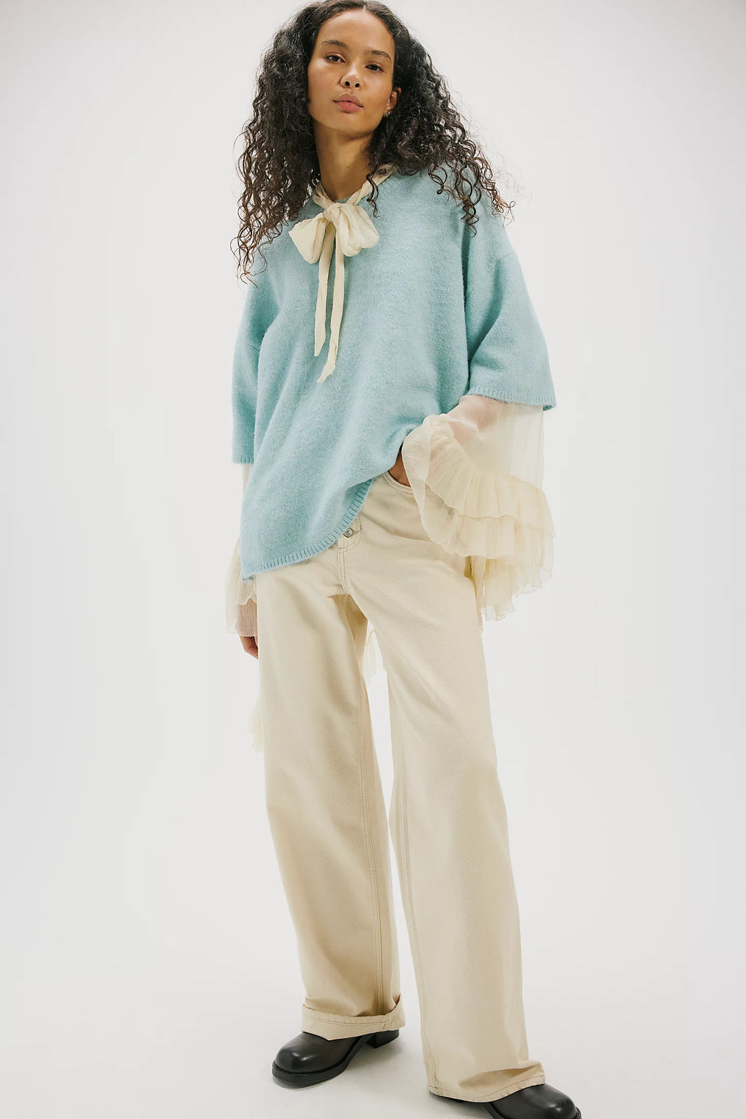 A model wearing a light blue knit sweater with a cream tie detail at the collar, layered over sheer cream ruffled sleeves, paired with cream wide-leg trousers and black shoes, against a plain white studio background.