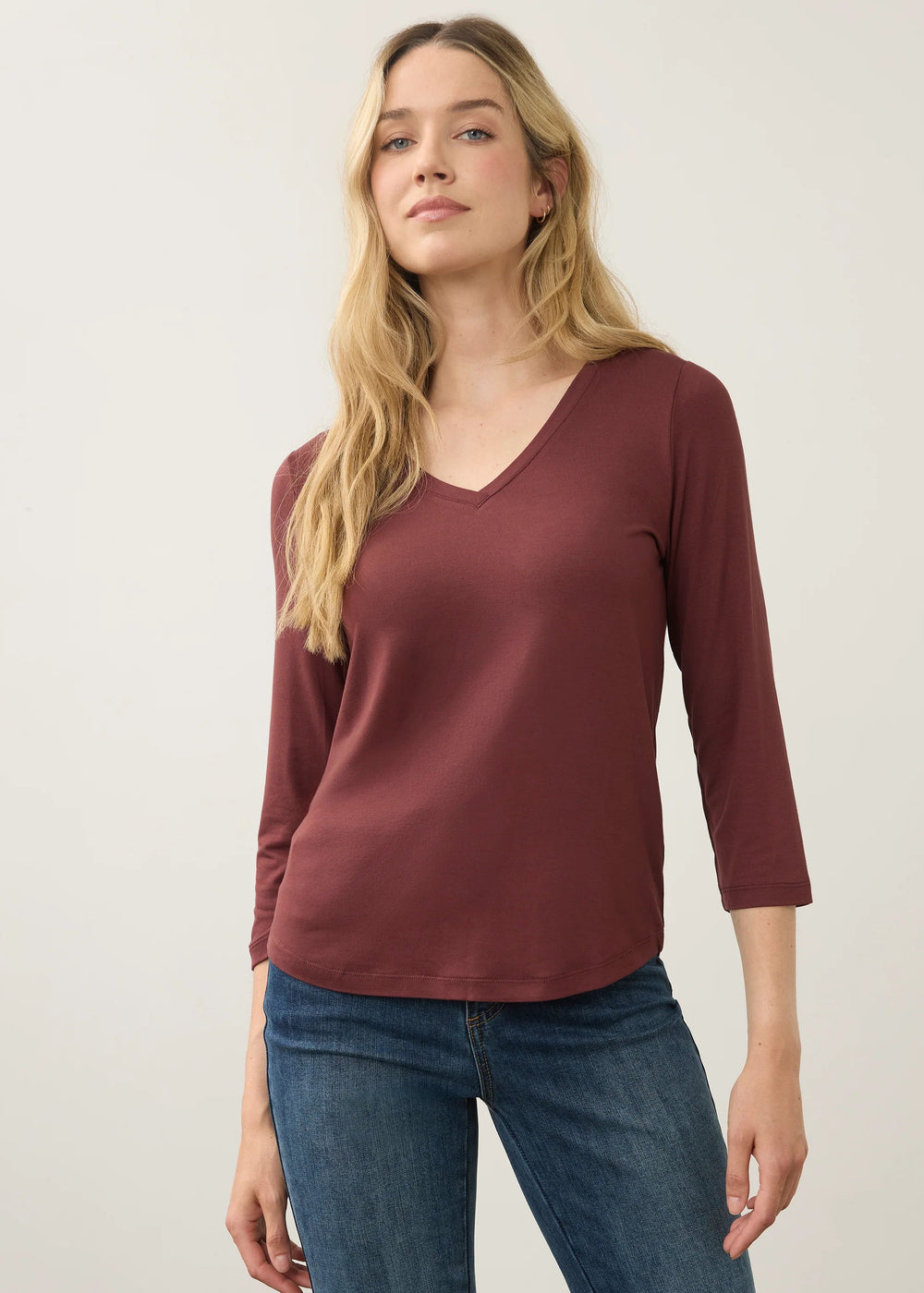 A woman modeling a three-quarter sleeve V-neck top in a dusty burgundy color, paired with blue jeans, against a plain light gray studio background.