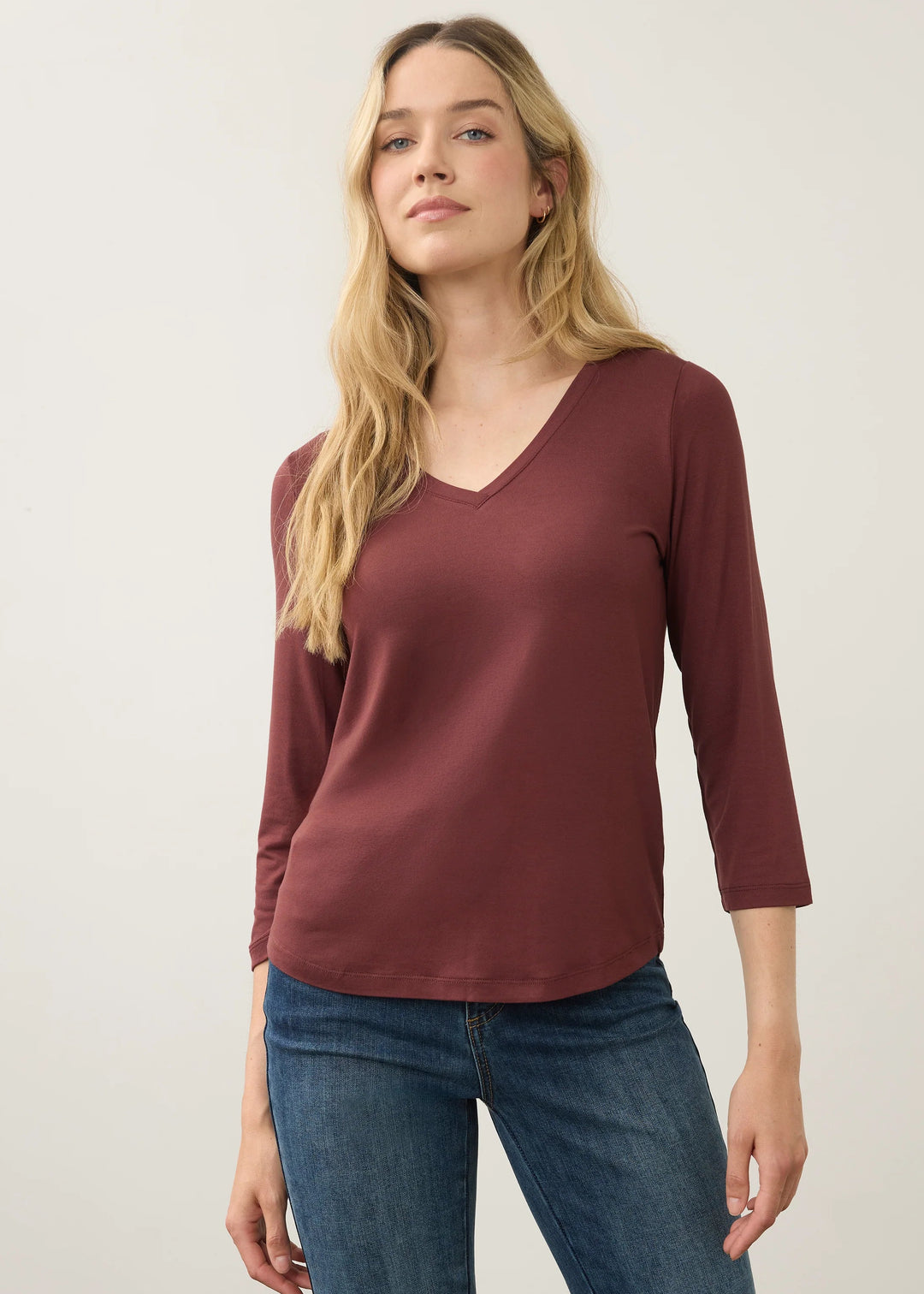 A woman modeling a three-quarter sleeve V-neck top in a dusty burgundy color, paired with blue jeans, against a plain light gray studio background.