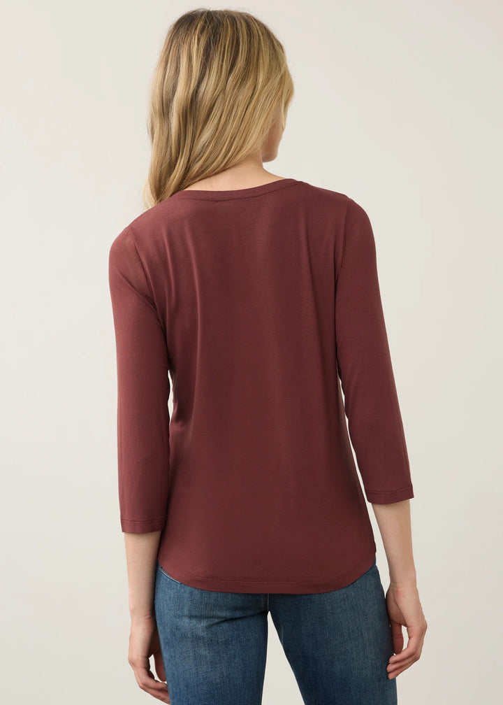 A woman viewed from the back wearing a rust-colored three-quarter sleeve top and blue jeans, standing against a plain off-white background.