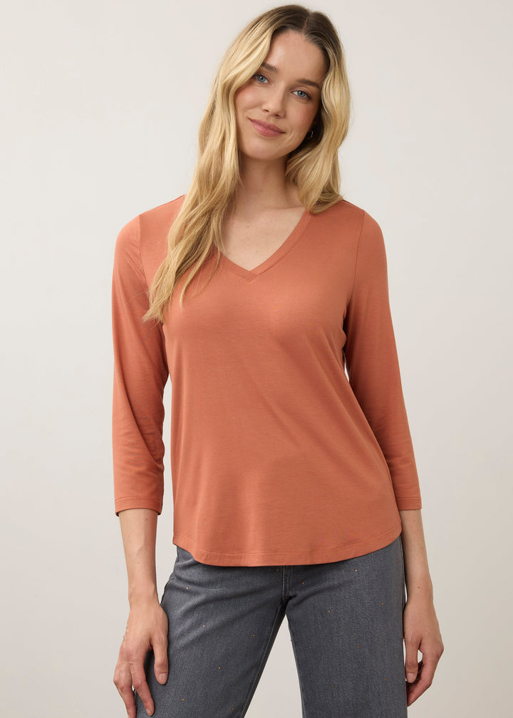 A woman modeling a three-quarter sleeve V-neck top in a muted terracotta or burnt orange color, paired with gray pants, against a plain light gray studio background.