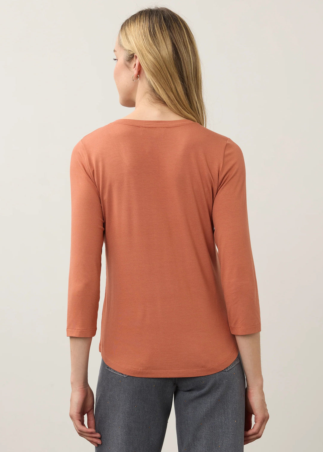 A woman shown from the back wearing a rust-orange three-quarter sleeve top and gray wide-leg pants, standing against a plain light gray studio background.