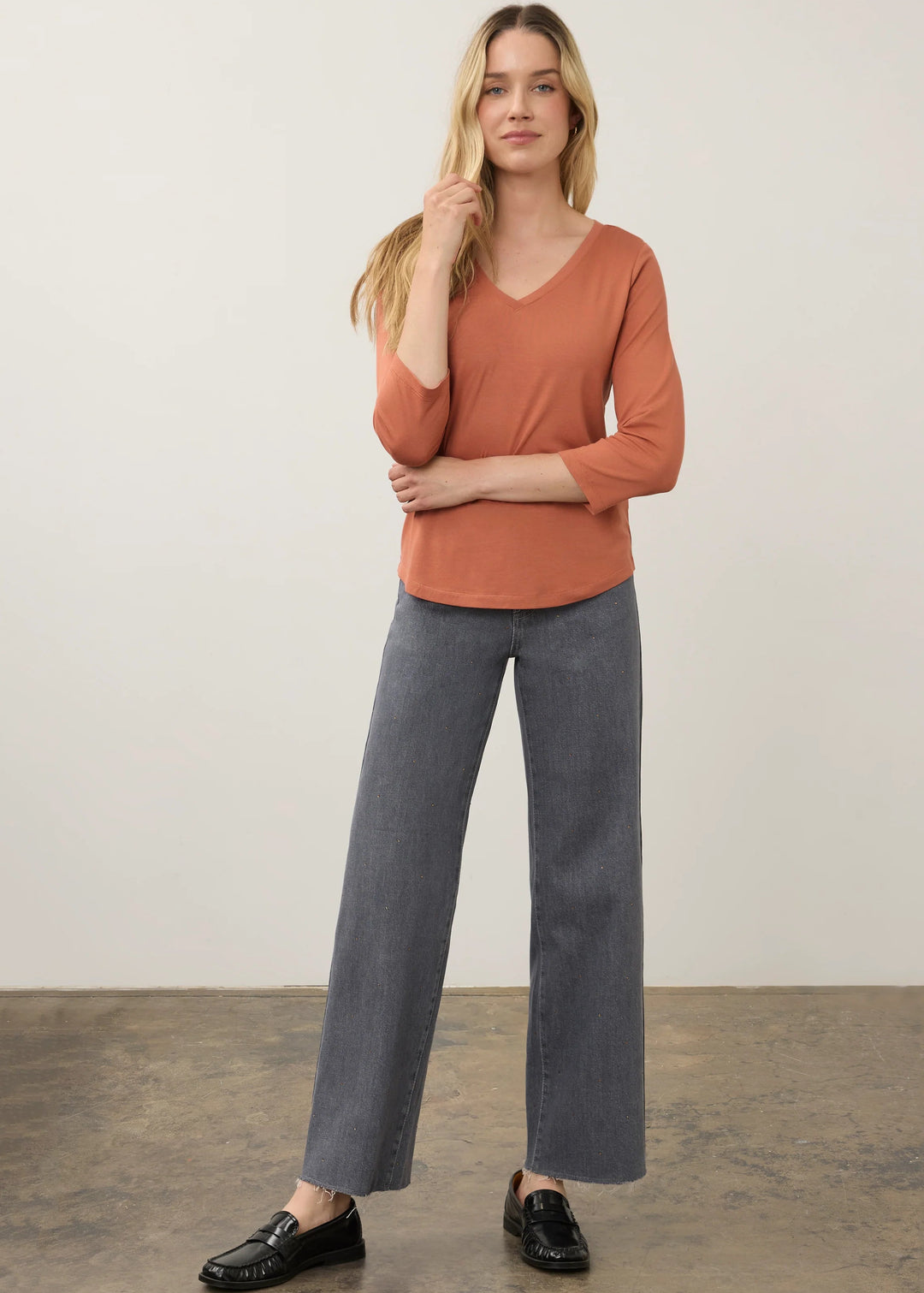 A woman modeling a rust-orange three-quarter sleeve V-neck top with wide-leg gray pants and black loafers, standing against a neutral studio backdrop.