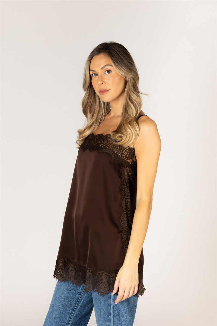 A woman wearing a dark brown satin camisole top with lace trim, paired with blue jeans, photographed against a plain light gray studio background.