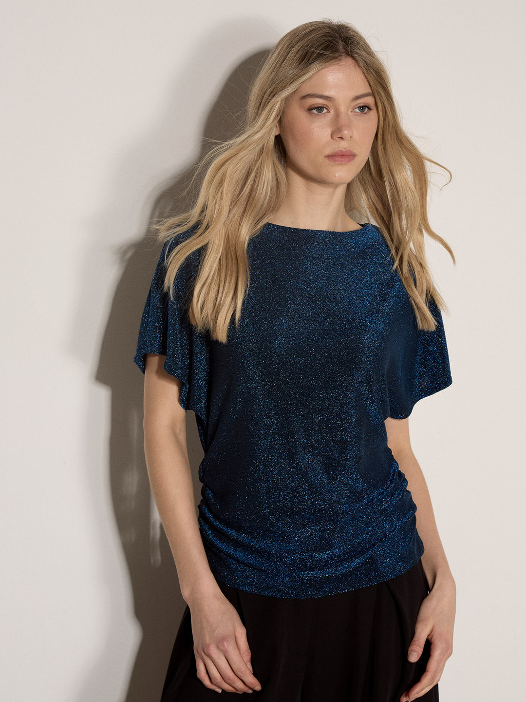A woman with blonde hair wearing a dark blue textured short-sleeve top and black bottoms, standing against a plain off-white wall.