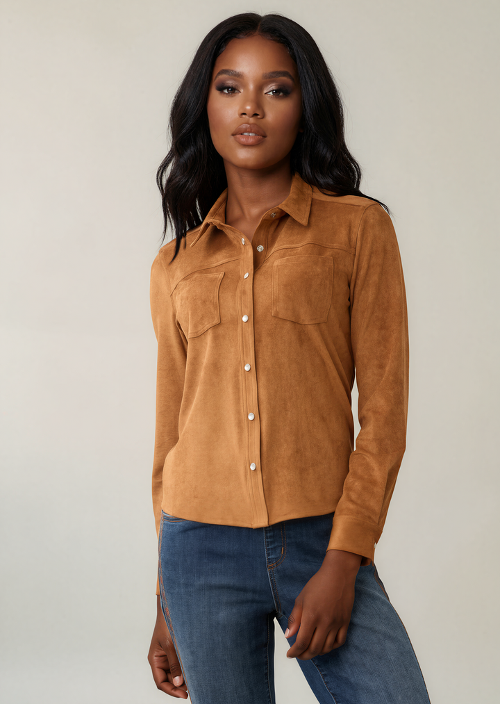 A woman modeling a tan suede long-sleeve button-up shirt with two chest pockets, paired with blue jeans, against a plain light beige studio background.
