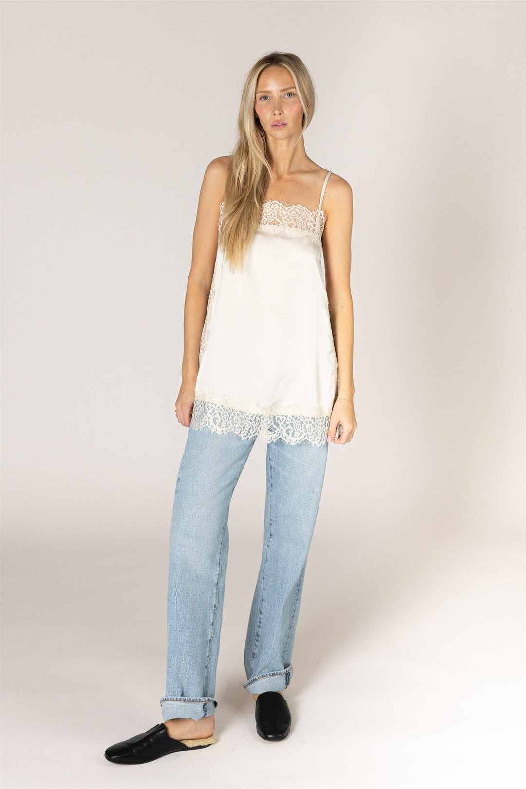 A woman modeling a white camisole top with delicate lace trim at the neckline and hem, paired with light-wash blue jeans and black flat shoes, against a plain light beige studio background.