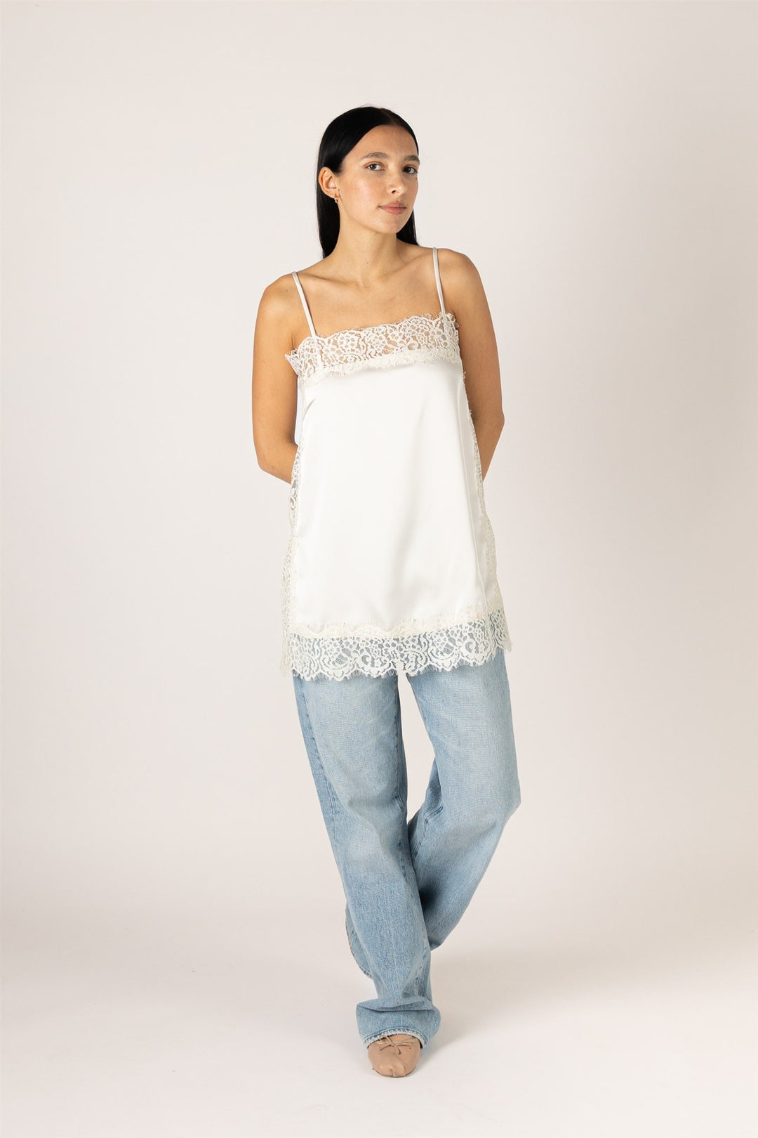 A woman modeling a white camisole top with delicate lace trim at the neckline and hem, paired with light-wash jeans, against a plain light beige studio background.