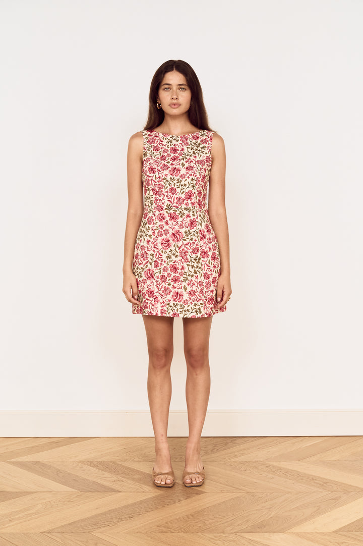 A woman stands in a sleeveless fit-and-flare mini dress with a pink, red, and green floral pattern on a cream background, paired with nude high-heeled sandals, set against a plain white wall and light wood herringbone floor.