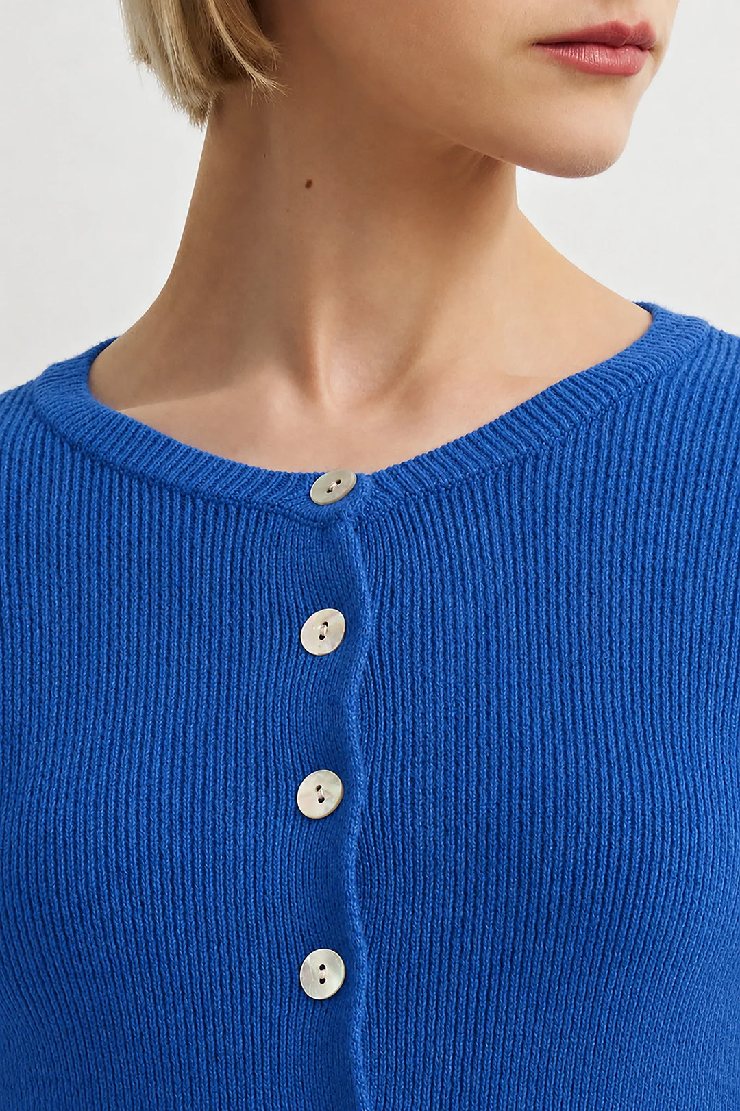 Close-up of a model wearing a vibrant blue ribbed knit cardigan with white buttons, showing the neck, jawline, and upper chest against a light gray background.