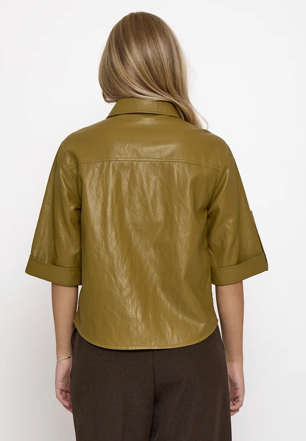 A woman viewed from the back wearing a cropped, short-sleeved olive green faux leather shirt and dark brown trousers, set against a plain white studio background.