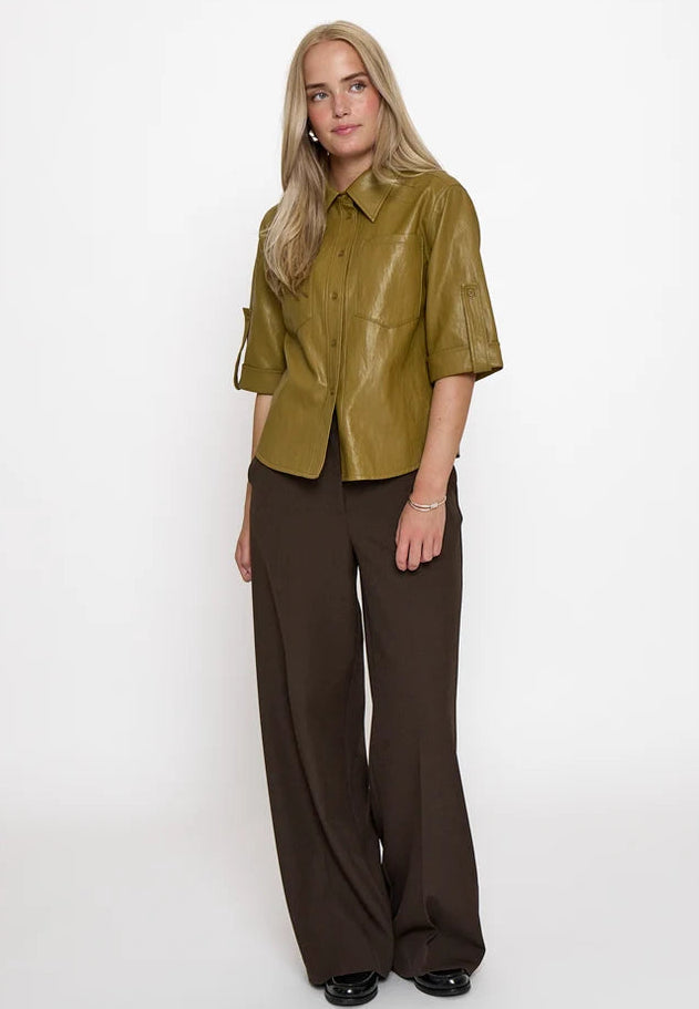 A blonde model stands against a plain white studio background wearing a short-sleeved olive green faux-leather button-up shirt and dark brown wide-leg trousers.