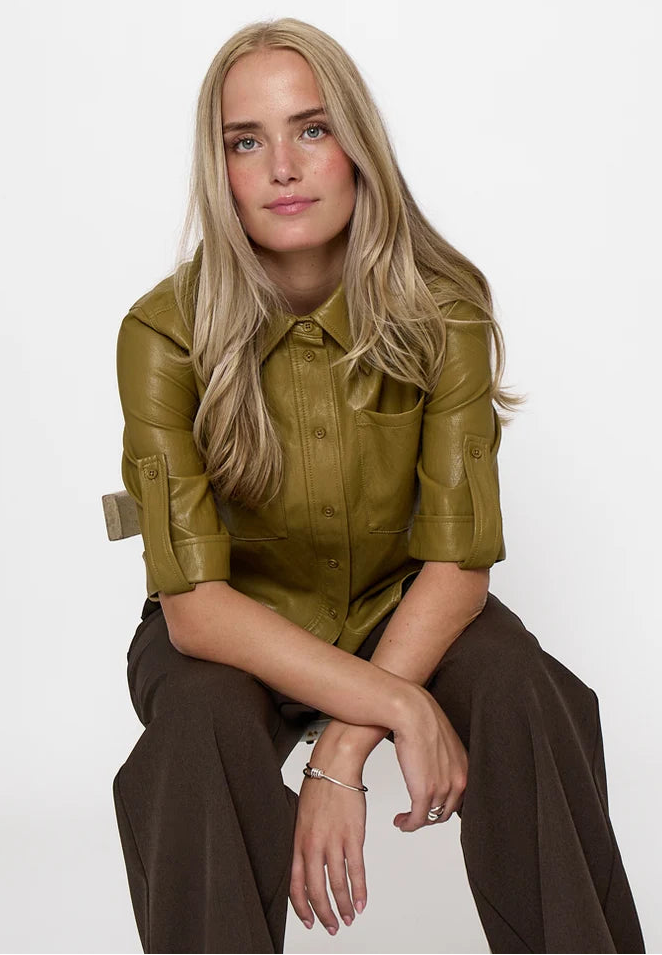 A blonde woman wearing a shiny olive green leather button-up shirt and dark brown wide-leg trousers, sitting against a plain white studio background.