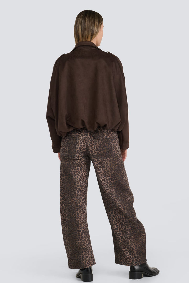 A model viewed from behind wearing a dark brown long-sleeve cropped jacket and brown leopard-print trousers, standing against a light gray studio background.