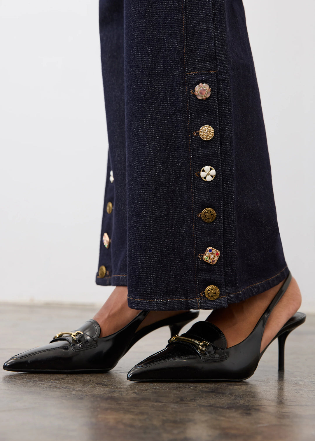 Close-up of a person wearing dark navy wide-leg jeans with decorative gold-tone buttons down the side and black pointed slingback heels with gold hardware, standing on a wooden floor against a light wall.