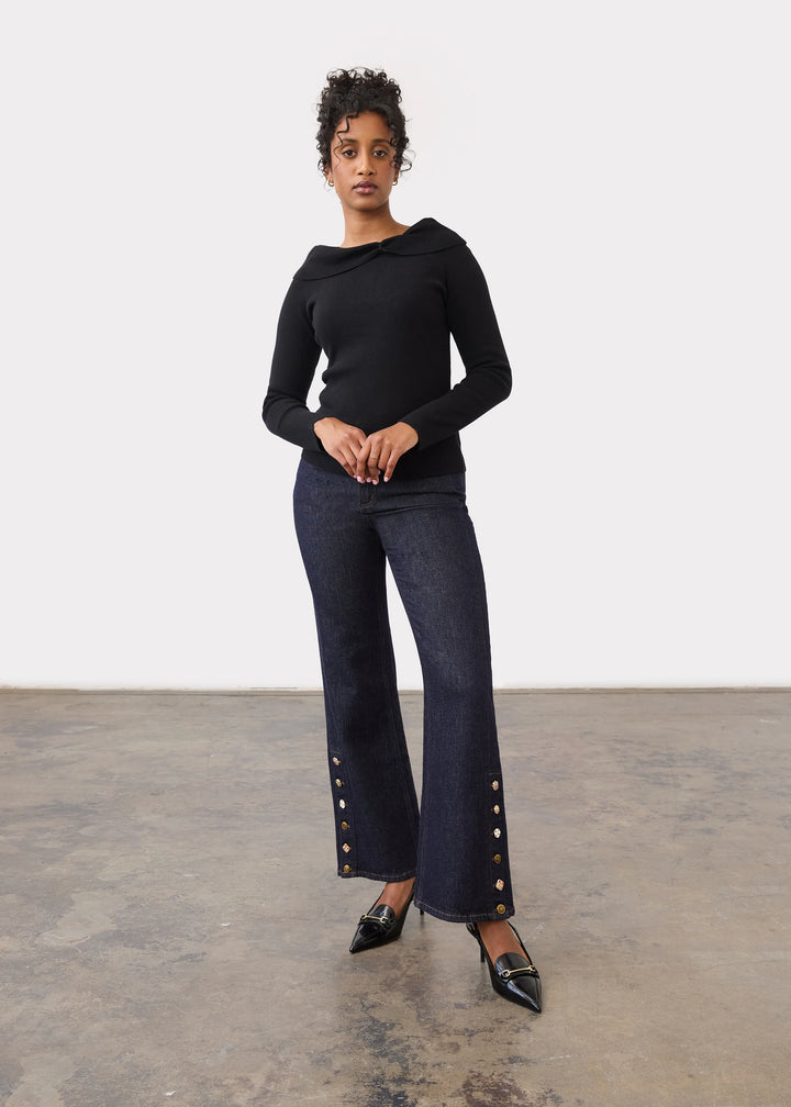 A model stands in a studio wearing a black long-sleeve top and dark blue wide-leg jeans with gold-tone button details near the hems, paired with black pointed shoes.