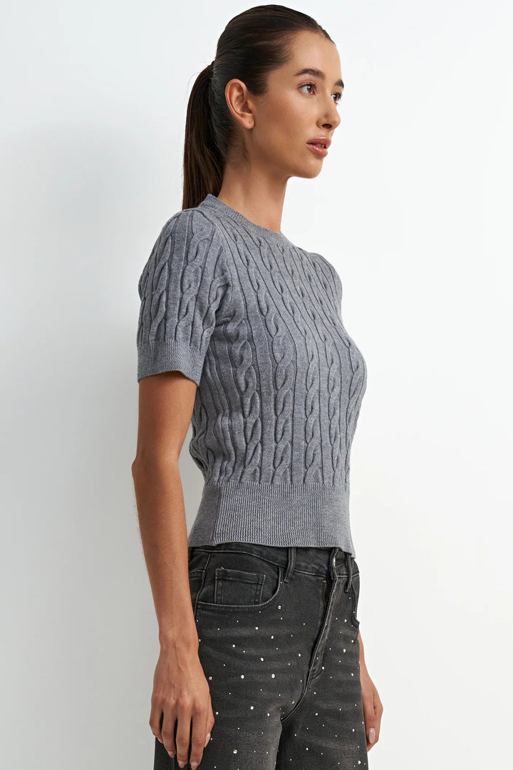 A woman wearing a gray short-sleeve cable-knit sweater and dark gray jeans with small white dots, standing against a plain light gray studio background.