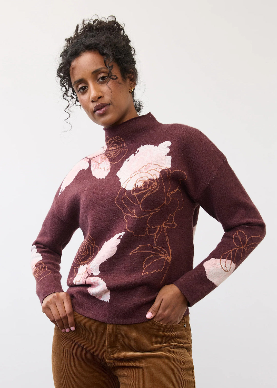 A woman modeling a burgundy mock-neck knit sweater with a pink and white floral pattern and brown corduroy pants, standing against a light gray studio background.