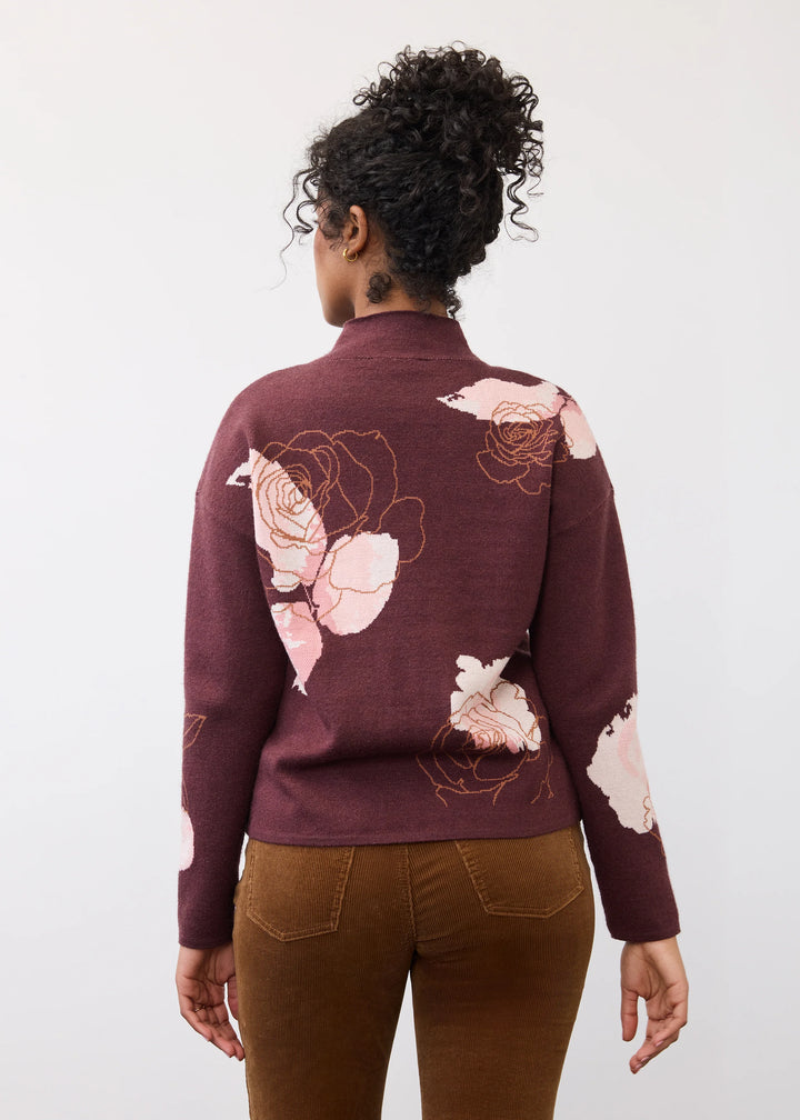 A woman viewed from behind wearing a burgundy mock-neck knit sweater with large pink rose patterns on the back and sleeves, paired with brown pants, against a plain white studio background.