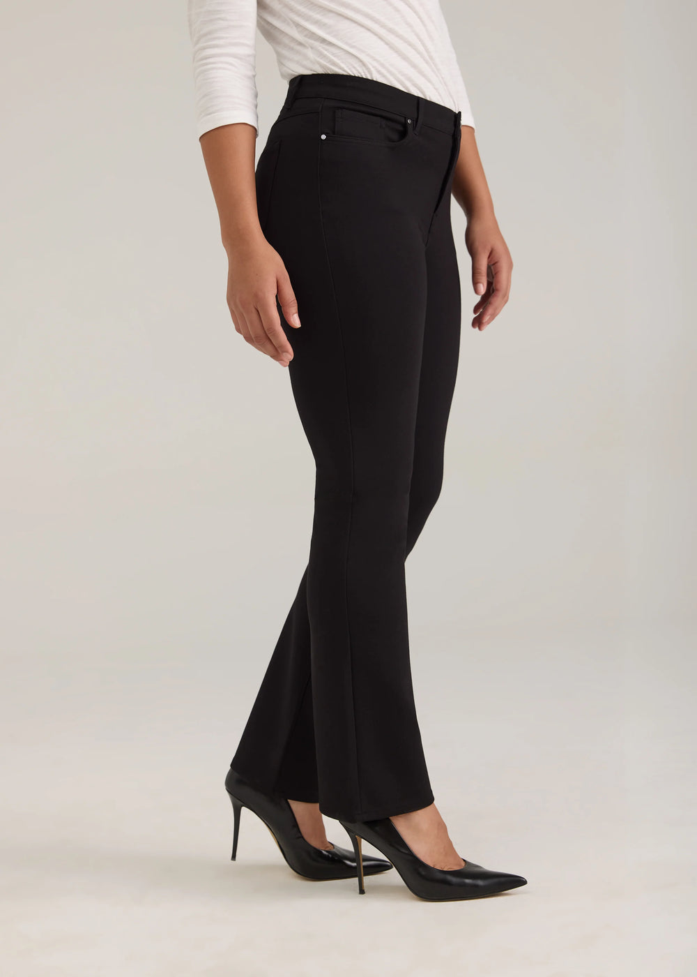 A model wearing black flared trousers, a white long-sleeve top, and black pointed-toe heels, shown from the waist down against a light gray studio background.