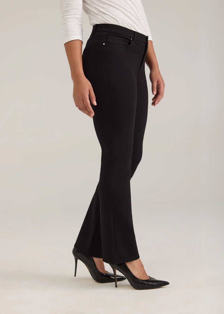A model wearing black flared trousers, a white long-sleeve top, and black pointed-toe heels, shown from the waist down against a light gray studio background.