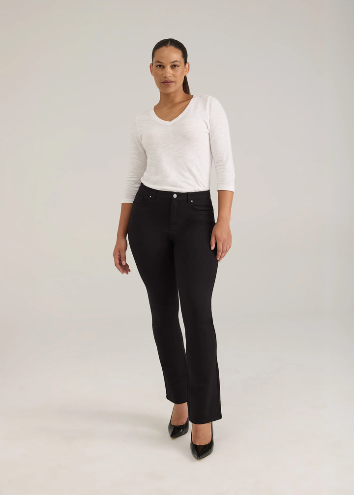 A woman modeling black high-waisted straight-leg jeans with a white long-sleeve top and black pointed-toe heels, standing against a light gray studio background.