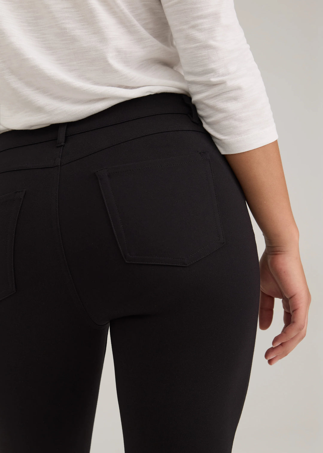 Close-up back view of a person wearing black high-waisted leggings with a back pocket, paired with a white long-sleeve top, against a light gray studio background.
