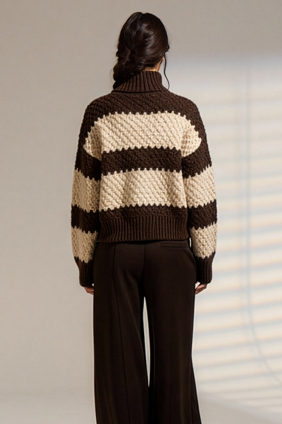 A model viewed from behind wearing a brown and cream striped chunky knit sweater and dark brown wide-leg trousers, standing against a light gray studio wall with soft window shadow patterns.