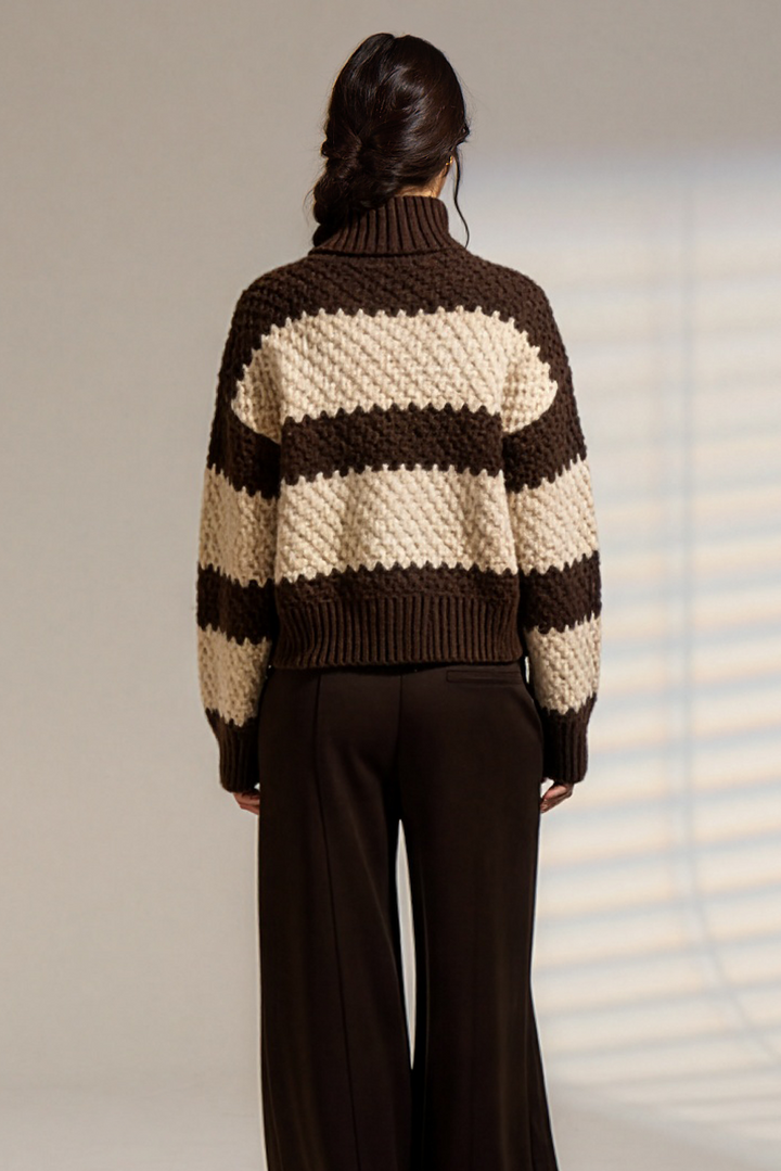 A model viewed from behind wearing a brown and cream striped chunky knit sweater and dark brown wide-leg trousers, standing against a light gray studio wall with soft window shadow patterns.