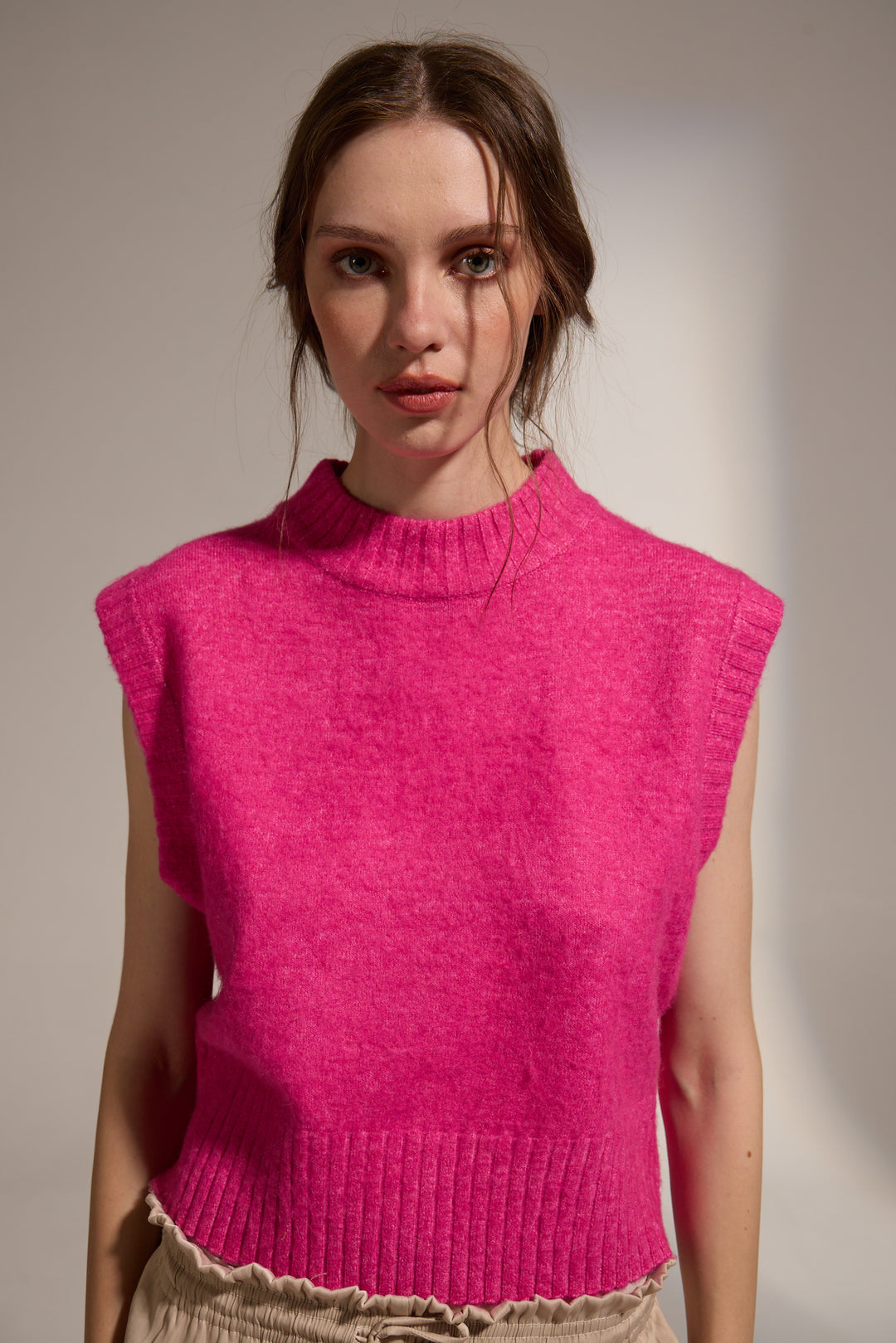 A woman wearing a sleeveless bright pink knit sweater vest with a ribbed mock neck and hem, paired with beige pants, photographed against a neutral studio background.